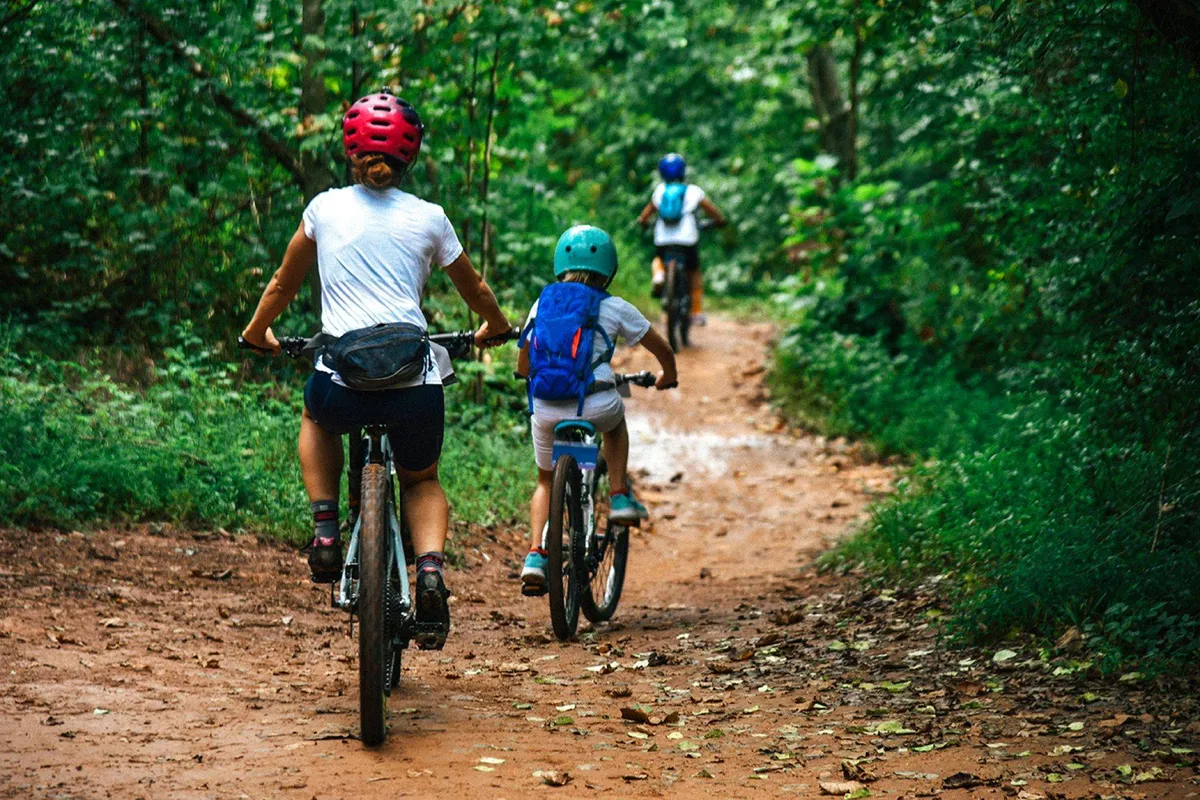 A lively family scene unfolds on a shaded dirt path, where bicyclists ride beneath a canopy of trees. The surroundings evoke energetic exploration and the joy of discovering nature together, with the possibility of connecting to local greenways or urban trails.