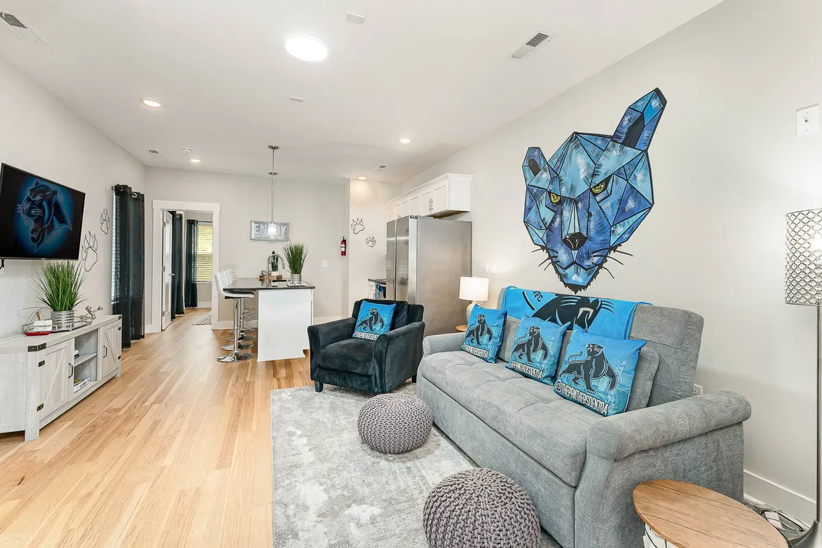 This image showcases a geometric wall art of a panther above a sofa adorned with decorative cushions. A modern light fixture hangs over the adjacent dining area, and a television is mounted on the wall. The space feels open and airy, with subtle playfulness from the art and a sophisticated layout that hints at a lively, inviting atmosphere.