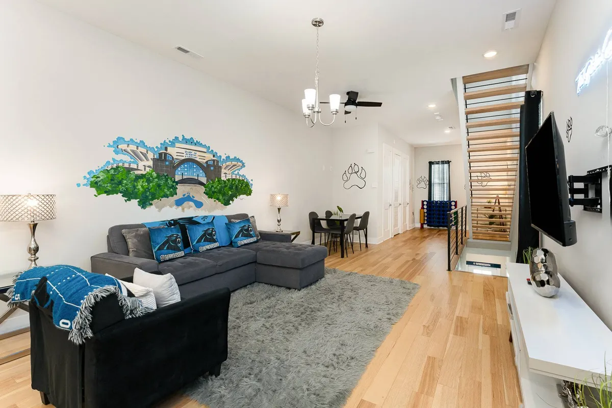 This image features a spacious, well-lit living and dining area with a modern sofa adorned with decorative pillows, a large artistic mural of a bridge that adds a local, urban flair, a sparkling chandelier, and a stylish dining table and chairs set up for group gatherings. The space feels comfortable and playful—perfect for entertaining friends or hosting celebratory meals.