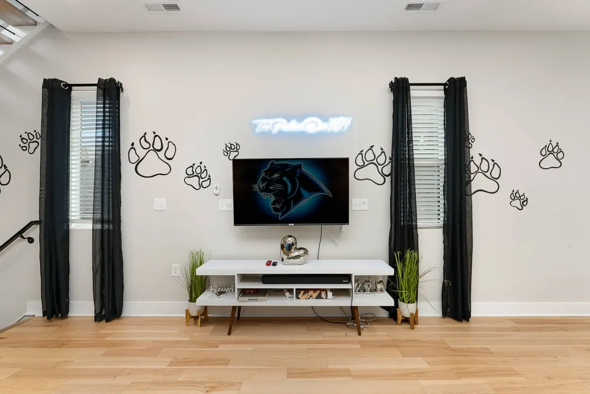 A bright, energetic living space featuring modern furniture, a TV with a vibrant panther graphic, and playful paw print wall decals. The setup encourages socializing, watching movies or games, and creating a lively, welcoming atmosphere for groups.