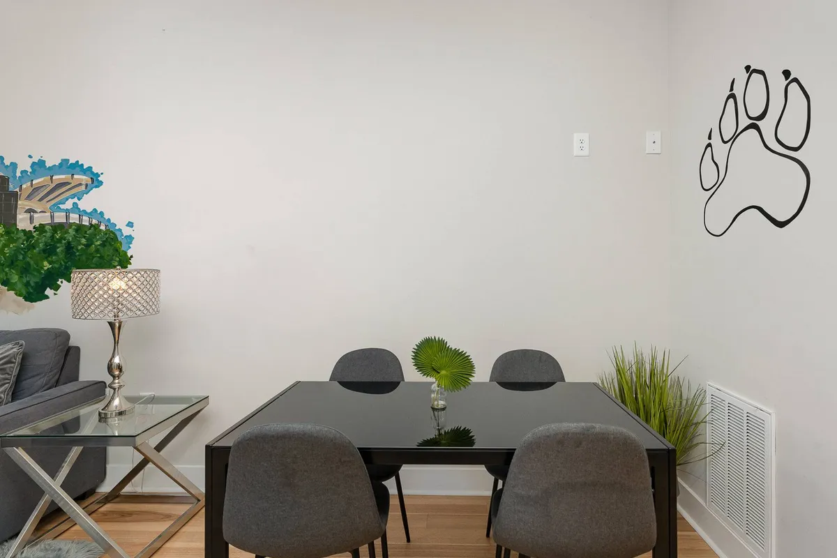 A modern, cozy dining area with a glass table, decorative lamp, green plant, and paw print wall art. The space is ideal for casual dining, social gatherings, or transforming into a flexible workspace for small group discussions, echoing the versatility needed for corporate retreats.