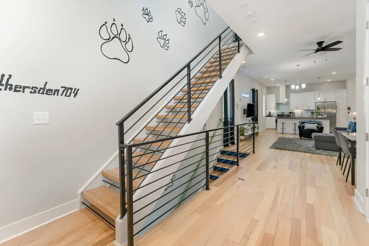 A spacious, open living area with a staircase featuring playful paw prints on the wall. The scene is lively, showing a group gathering space where guests can relax, socialize, or enjoy a meal in the adjoining bright kitchen. The arrangement invites guests to feel at home and signals a pet-friendly environment.