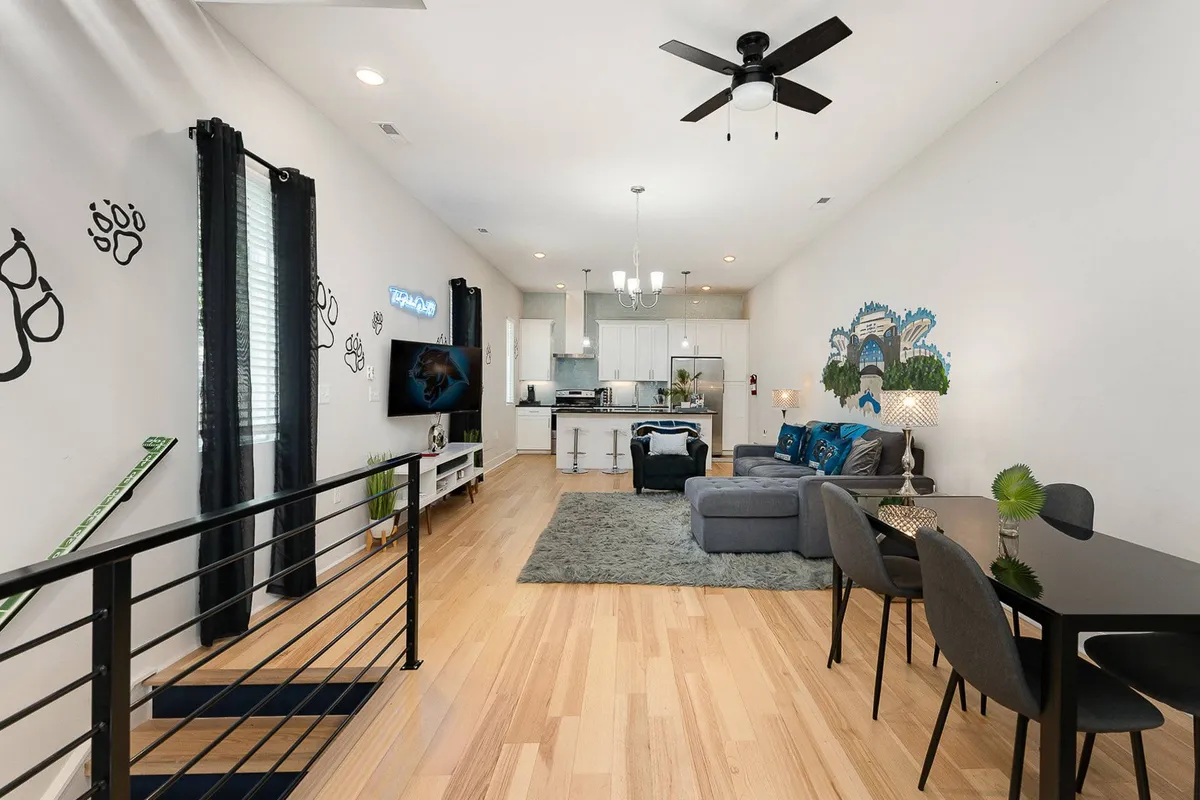 The photo captures a comfortable living area with a plush sofa, a dining table positioned for group interactions, a TV for entertainment, and eye-catching wall art. The overall mood is lively and welcoming, with large windows that allow natural light to pour in.